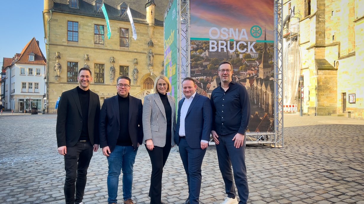 Gruppe steht vor den Plakaten und dem beflaggten Rathaus mit dem neuen Osnabrück-Logo